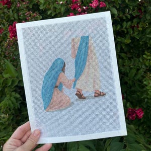 May include: A needlepoint canvas featuring a printed design of a kneeling figure in a tan garment with a blue head covering, reaching toward a standing figure in a tan robe with a blue sash and brown sandals, all on a white grid background.