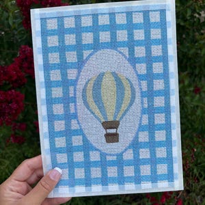 May include: A rectangular needlepoint canvas featuring a blue and white gingham check pattern. Centered on the canvas is a white oval containing a graphic of a hot air balloon with yellow and blue vertical stripes and a brown basket.