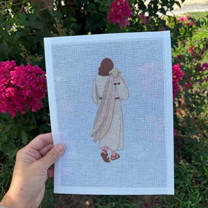 May include: A rectangular needlepoint canvas featuring a cross-stitched figure viewed from behind, wearing a long beige robe and brown sandals, carrying a small white lamb over the shoulders, set against a white grid background.