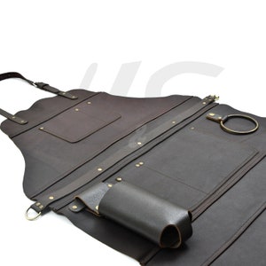 Può includere: Un grembiule in pelle marrone con più tasche, un portautensili e un anello di metallo. Il grembiule ha spalline regolabili ed è fissato con rivetti in ottone.