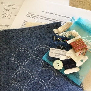 May include: A Sashiko embroidery kit featuring a piece of blue denim fabric with a white arched pattern, a light blue organza bag, sewing needles, a white button, and three spools of thread in pink, white, and dark blue, with printed instructions.