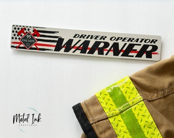 May include: A rectangular sign with the text "DRIVER OPERATOR WARNER" in black, with a red line through the name. The sign features a red and black American flag design with crossed axes and the text "95 FIRE DEPT. ESTD. 1995".
