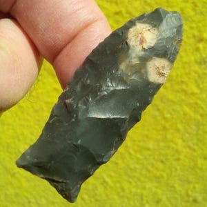 May include: A dark gray, chipped stone arrowhead with two beige patches. The arrowhead is held between fingers, showcasing its sharp edges and textured surface. The stone appears to be obsidian or a similar material, with a slightly glossy finish.