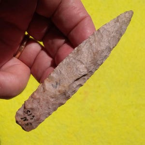 May include: A hand-held, light brown, stone arrowhead with a sharp point. The arrowhead has a rough texture and appears to be hand-chipped. The base of the arrowhead has the markings "SN 5.15".
