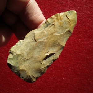 May include: A light brown, arrowhead-shaped stone tool with a rough, textured surface. The tool is held in a hand against a red background.