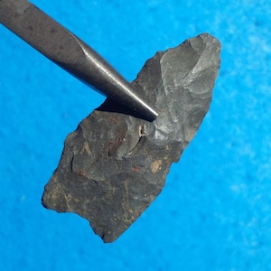 May include: A dark gray, chipped stone arrowhead with a pointed tip and a notched base. The arrowhead features a textured, flaked surface and is held by a metallic tool. The object has a distinct triangular shape with serrated edges along the sides.