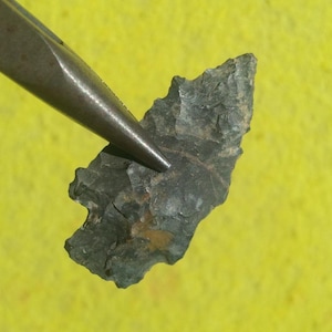 May include: A dark gray, chipped stone arrowhead held by a pair of metal pliers. The arrowhead features a pointed tip, serrated edges, and a notched base, with visible flaking patterns and a small patch of brown discoloration on its surface.