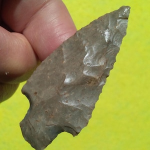 May include: A gray, triangular stone arrowhead featuring a chipped, textured surface with a pointed tip and a notched base. The object displays a series of small, irregular indentations along its edges, characteristic of a hand-knapped stone tool.