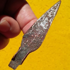 Puede incluir: Una punta de flecha metálica desgastada con una punta afilada y una superficie texturizada. La punta de flecha tiene un tono marrón rojizo con reflejos plateados y una base ahuecada. El fondo es amarillo liso.