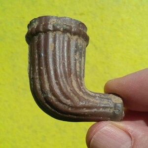 May include: A brown, L-shaped tobacco pipe bowl featuring a vertical ribbed texture along its curved body. The pipe has a wide, circular opening at the top and a smaller, cylindrical stem end, showing signs of wear and a weathered, matte finish.