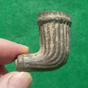 Puede incluir: Una pequeña cazoleta de pipa de tabaco en forma de L, de color marrón oscuro, con una textura acanalada vertical en la superficie exterior. El borde superior de la cazoleta está decorado con un sutil patrón de cuentas, con aspecto envejecido.