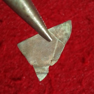 Può includere: Primo piano di un pezzo di rame rotto, forse una moneta, con una patina verdastra. Il rame è tenuto da una pinza.