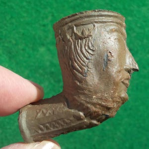 May include: A brown, antique tobacco pipe shaped like a human head. The pipe features detailed carvings of hair, facial features, and decorative patterns. The pipe is held by a person's finger.