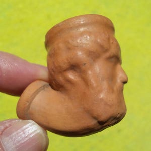 May include: A terracotta-colored clay pipe bowl featuring a molded relief of a human face. The object has a smooth, matte finish with a cylindrical opening at the top and a short, protruding stem base extending from the side of the sculpted head.