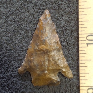 May include: A brown arrowhead with a pointed tip and a slightly curved base. The arrowhead is about 1 inch long and is resting on a dark gray surface. A ruler is visible on the right side of the image.