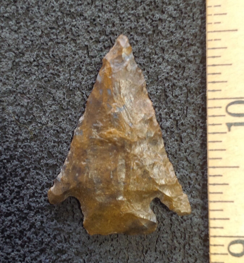 May include: A brown arrowhead with a pointed tip and a slightly curved base. The arrowhead is about 1 inch long and is resting on a dark gray surface. A ruler is visible on the right side of the image.