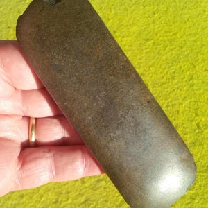 Puede incluir: Un artefacto de piedra alargado de color gris oscuro, con un extremo redondeado y una superficie rugosa y texturizada. La piedra se sostiene en una mano, mostrando su tamaño y forma. El objeto parece ser una herramienta antigua o una obra de arte primitiva.