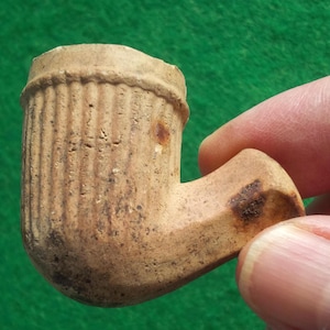 May include: A brown clay pipe with a ribbed bowl and a curved stem. The pipe is slightly worn and has a few brown spots.