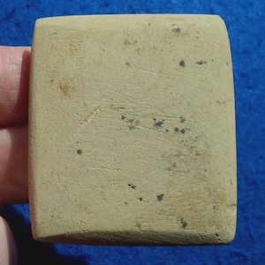 May include: A rectangular, beige stone with rounded corners. The stone has a slightly textured surface with small dark spots and markings. The object is held by a hand, with a blue background visible.
