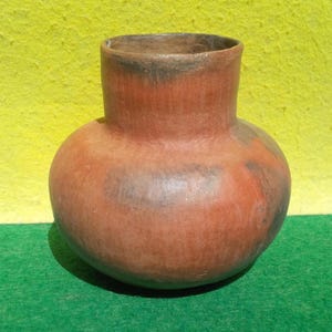 May include: A round, terracotta-colored clay pot with a wide, bulbous body and a short, cylindrical neck. The surface features natural variations in color, including patches of dark gray and black, with a matte, unglazed, and textured finish.