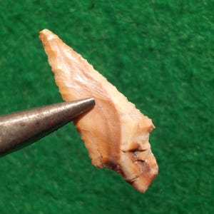 May include: A small, triangular, arrowhead made of brown and white banded stone. The arrowhead is about 2 inches long and has a sharp point.
