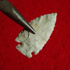 Peut inclure: Une pointe de flèche en pierre claire et ébréchée, avec une base crantée, possiblement un artefact ancien. Un outil en métal est positionné près de la pointe de flèche. Le fond est rouge uni.