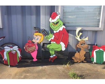 May include: A Christmas yard decoration set featuring the Grinch, Cindy Lou Who, and Max the dog. The Grinch is wearing a red Santa suit and a green hat. Cindy Lou Who is holding a pink Christmas ornament with the name "Jaynes" on it. Max the dog is wearing a reindeer antler headband. The set also includes two green and red gift boxes.