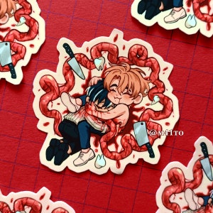 Puede incluir: Una pegatina con dos figuras de dibujos animados abrazándose, rodeadas de tubos rojos y carnosos y salpicaduras de sangre. También hay cuchillos y dientes. La pegatina tiene un borde blanco y está sobre un fondo de cuadrícula roja. El texto "@M41TO" es visible.