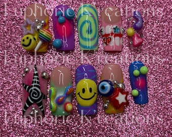 May include: A set of ten artificial nails with various designs. The nails feature a range of colors, including pink, purple, blue, and green, and are adorned with embellishments such as smiley faces, stars, and swirls. Some nails have 3D elements.