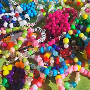 May include: A collection of colourful beaded bracelets with various letters and symbols. The bracelets are made of plastic beads and are strung together in different patterns. Some of the bracelets have words spelled out on them, such as "PIG", "SA", "WSER", "UK", "URS", "PE".