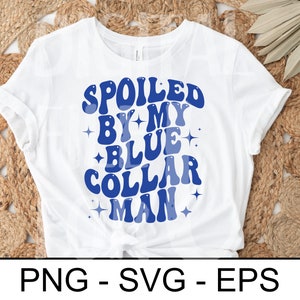 以下が含まれることがあります： レトロなフォントで「SPOILED BY MY BLUE COLLAR MAN」と青い文字がプリントされた白いTシャツ。星のアクセント付き。織り込まれたニュートラルな色調の表面にシャツが置かれています。
