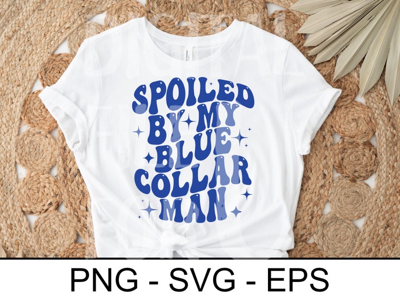 以下が含まれることがあります： レトロなフォントで「SPOILED BY MY BLUE COLLAR MAN」と青い文字がプリントされた白いTシャツ。星のアクセント付き。織り込まれたニュートラルな色調の表面にシャツが置かれています。