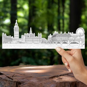K&ouml;nnte beinhalten: Eine Schwarzwei&szlig;-Zeichnung der Londoner Skyline mit Big Ben, dem Houses of Parliament und dem London Eye. Die Zeichnung wird in einer Hand vor einem verschwommenen gr&uuml;nen Hintergrund gehalten.