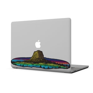 May include: A laptop computer with a colorful sticker of a mountain range and a sunset. The sticker is on the bottom of the laptop and features a stylized depiction of a mountain range with a sunset in the background. The sticker is in shades of blue, green, purple, and orange.