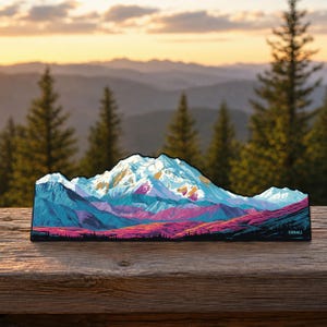 Puede incluir: Un letrero decorativo plano y troquelado con una ilustración estilizada de la montaña Denali. La obra muestra picos nevados en blanco y azul, con tonos vibrantes de morado, rosa y rojo en las laderas inferiores. La palabra DENALI está impresa.