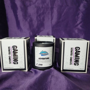 May include: Three black candle jars with white labels. The center jar has a label that says "Adventure" and features a mountain range graphic. The labels on the other two jars say "Gaming Scented Candles".
