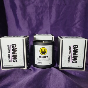 Può includere: Tre scatole nere con testo bianco che recita "Gaming Scented Candles" e una candela nera con un'etichetta bianca che recita "Comedy" e una grafica di un viso sorridente. La candela ha una descrizione del profumo "Miscela di banana, pompelmo, sempreverde, cocco, arancia, vaniglia e lampone. È un'allure cremosa di vaniglia e chewing gum."