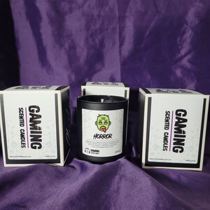 May include: Three black boxes with white labels that say "Gaming Scented Candles" and a black candle with a white label that says "Horror" and a cartoon green zombie with a tongue sticking out. The candle is labeled "Gaming Scented Candles".