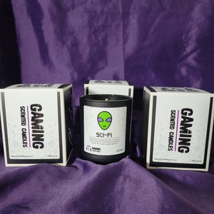 May include: Three black boxes with white text that reads "Gaming Scented Candles" and a black candle with a white label that reads "Sci-Fi" with an alien graphic. The candle has a white floral fragrance with sweet amber, balsamic notes, precious woods and exotic Indian Jasmine.