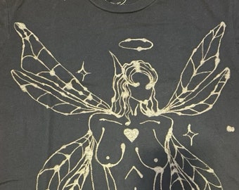 May include: A dark gray t-shirt with a gold-colored print of a winged figure with a halo. The figure has a heart on its chest and is surrounded by stars.