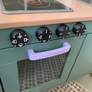 Könnte beinhalten: Ein türkisfarbener Spielofen mit schwarzem Kochfeld und vier schwarzen Knöpfen. Die Knöpfe haben weiße Markierungen und Zahlen. Die Backofentür hat einen lilafarbenen Griff mit dem Text "hier kocht" in weiß.