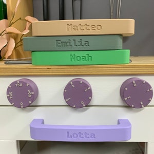Può includere: Un fornello giocattolo bianco con manopole viola e una maniglia viola. La maniglia ha il nome "Lotta" inciso. Tre blocchi rettangolari con i nomi "Matteo", "Emilia" e "Noah" sono impilati sopra il fornello.