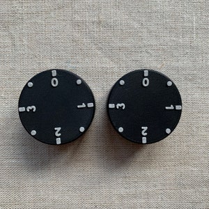 May include: Two black circular dials with white numbers and markers. Each dial has the numbers 0, 1, 2, and 3, along with small white dots. The dials are set against a neutral, textured background.