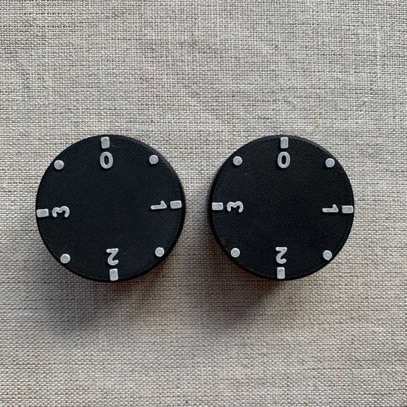 May include: Two black circular dials with white numbers and markers. Each dial has the numbers 0, 1, 2, and 3, along with small white dots. The dials are set against a neutral, textured background.