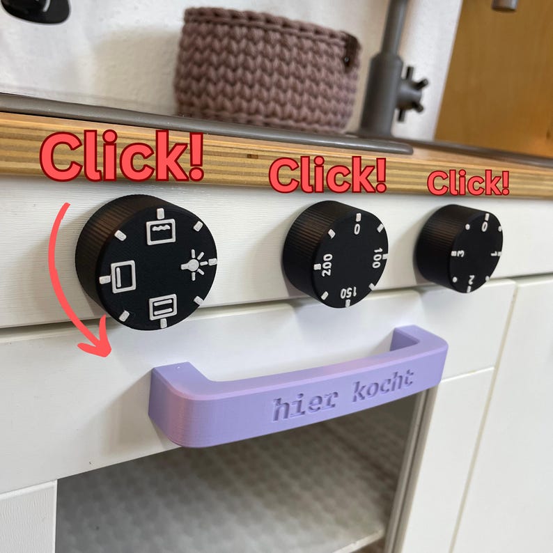 May include: A close-up of a toy kitchen appliance with black knobs and a light purple handle. The knobs have white symbols and numbers. The handle has the text "hier kocht". The word "Click!" is written above the knobs.