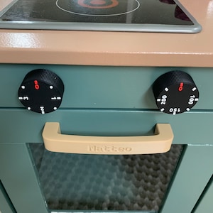 Może przedstawiać: Zabawkowy piekarnik w kolorze turkusowym z czarną płytą grzewczą, dwoma czarnymi pokrętłami i beżowym uchwytem z nadrukowanym napisem "Matteo".