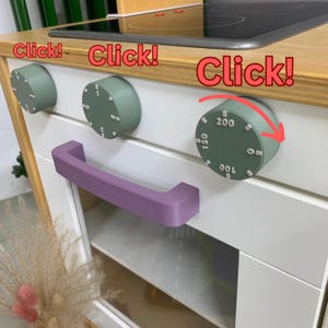 Op de afbeelding: Een speelkeuken met een witte kast, houten aanrechtblad en een paarse handgreep. Het heeft groene knoppen met cijfers en het woord "Click!" in het rood.