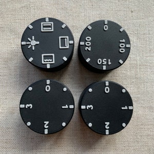 May include: Four black control knobs with white markings. Two knobs have symbols, one with a lightbulb, a rectangle, and a wave. The other two knobs have numbers, including 100, 150, and 200. The other two knobs have numbers 0, 1, 2, and 3.