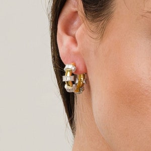 May include: Gold hoop earrings with rectangular and round-cut clear stones. The earrings have a two-tone design, with a gold-coloured metal and clear stones. The earrings are worn on a person's ear.