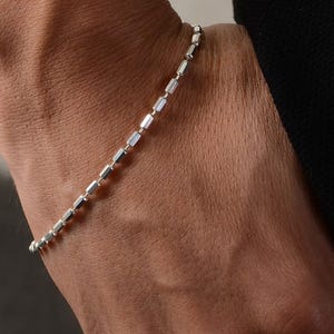 May include: A silver chain bracelet with a delicate, linked design. The bracelet is worn on a person's wrist.